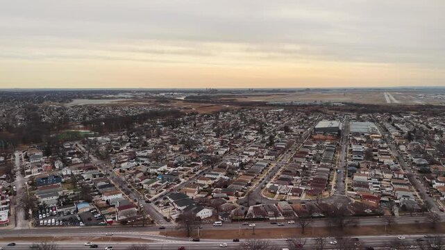 A drone pans across a suburban street grid in Queens beneath pale clouds. Closely spaced houses and parallel roads form a dense residential pattern with bare trees and winter light to the horizon.