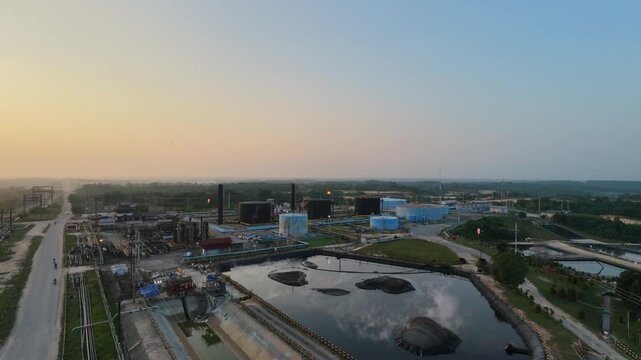 Aerial view of an industrial oil refinery plant with large storage tanks, piping systems, and processing facilities under a glowing sunset sky. Energy, fuel production, and industry concepts
