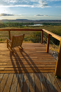 A rocking chair overlooking a river casts long shadows on a wood deck at dusk