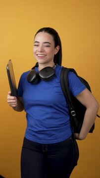 Happy latina female university student dancing with headphones, backpack, and laptop on a yellow background