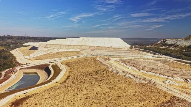 Terraced excavation and water ponds cover an open-pit mining site, with exposed earth and distant hills under a blue sky during the day.