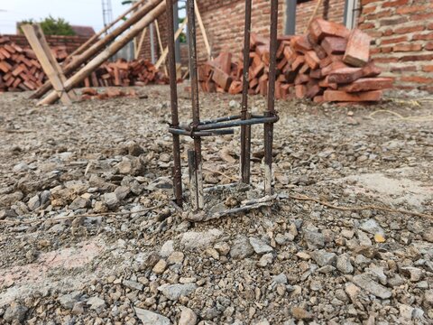 Steel rebar column reinforcement emerging from gravel ground on active house construction site with red bricks pile in background, ideal for civil engineering and structural building photo.