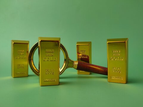 A brass magnifying glass with a wooden handle rests in front of four upright gold bullion bars on green