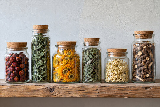 Row of corked glass apothecary jars filled with dried herbs, calendula flowers, rose hips and licorice root on a rustic wooden shelf