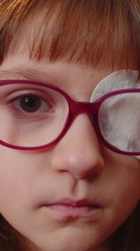 Sick ill toddler girl face looking at camera having protective bandage plaster patch scratch on one eye. Injury trauma blind. Child kid wearing glasses at home room. Medical eyesight vision therapy