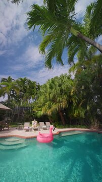 Vertical Screen: Woman relaxing by the pool on a sun lounger in a white hat. Pink flamingo float on turquoise water with a brick edge. Lush palm garden and blue sky with clouds. Sunny summer vacation.