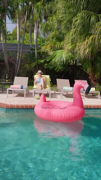 Vertical Screen: Woman relaxing by pool scrolls her phone and holds an orange cocktail on a lounger. Pink flamingo float on turquoise water. Champagne bottle on a side table. Palm trees on a sunny day