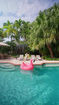 Vertical Screen: Woman relaxing by pool sips cocktail on a yellow towel lounger in a sunny backyard. Pink flamingo float on turquoise water, tall palm trees, patio table and umbrella. Summer vacation.
