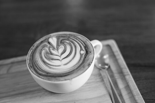 Black and white coffee cup with latte art on wooden tray beside spoon, creating calm cafe setting with monochrome texture and warm beverage detail