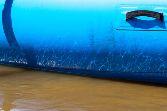 Blue inflatable raft floating on muddy river water with sunlight reflections and black carrying handle, suggesting outdoor recreation and water travel