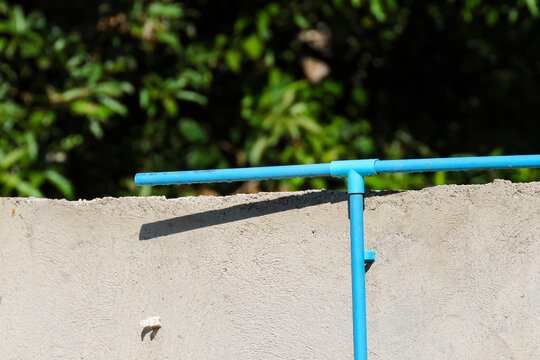 Blue water pipe above rough concrete wall with green foliage and strong sunlight creating clear diagonal shadow, representing outdoor plumbing and utility infrastructure in residential setting