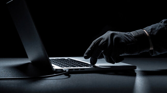 Gloved hand uses laptop on dark desk. Hacker types on keyboard in low key lighting. Cyber crime concept shows unauthorized access to computer data. Criminal inserts usb drive to steal private info