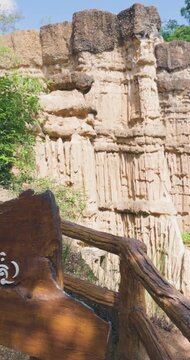 Vertical pan leaves wooden PHA CHOR sign to reveal layered earth towers, tree foliage and railed path at Pha Chor, Mae Wang National Park, Doi Lo, Chiang Mai, Thailand. Signage introduces canyon.