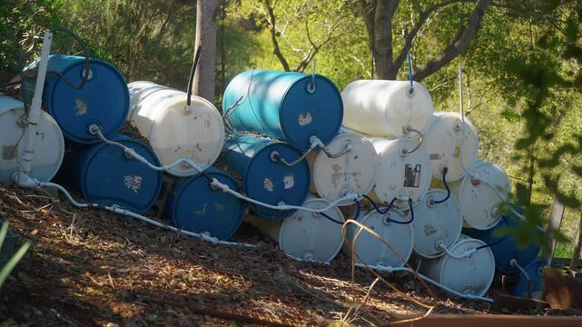Array Of Connected Blue And White Plastic Storage Barrels Stacked On Hillside Slopes For DIY Off Grid Rainwater Catchment System. - wide shot