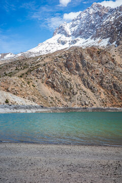 Beautiful alpine landscape featuring a turquoise mountain lake with a gravel shoreline, surrounded by rugged rocky cliffs and towering snow capped peaks