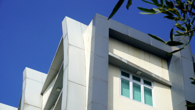 White curved concrete facade with cream wall and green curtained windows, framed by leafy branch at upper right, rising against clear deep blue sky in low angle view.