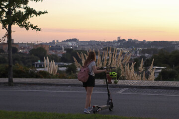 Young woman with backpack crossing a road with an electric scooter at sunset, city view and pampas grass in the background. Suitable for urban mobility, eco transport, lifestyle and travel themes. © Evgeniia