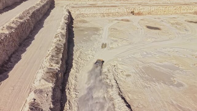 During daytime, an earthmoving truck drives through a quarry, raising dust beside deep excavated walls and tracks in the ground.