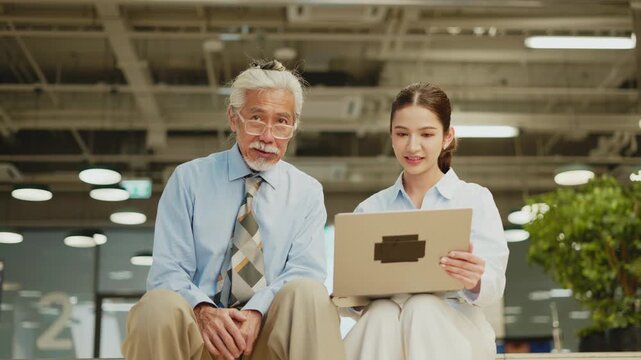 Young Thai woman holding laptop explains to smiling senior man