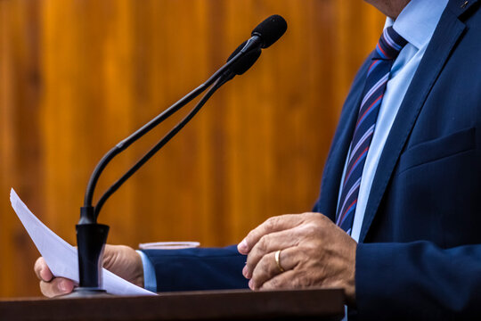 Close-up of a businessman or politician wearing a green suit and watch gesturing during a speech with microphones at a podium. Detail of a speaker's hands and microphones 