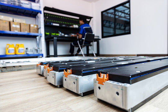 Electric vehicle battery modules arranged in the foreground of a battery inspection workshop, with a technician seated at a workbench in the background. 