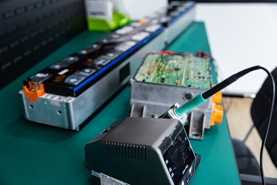 Close-up of testing equipment and a wired probe on a workbench, with electric vehicle battery modules and an exposed circuit board in the background. 