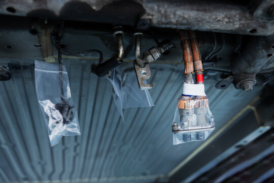 Close-up of a disconnected electric vehicle battery connector and orange cables beneath a car, with transparent wrapping around the connector during automotive maintenance.