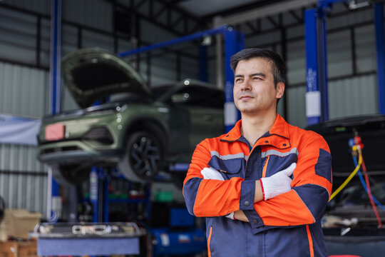 Confident electric vehicle mechanic wearing orange and navy workwear and protective gloves stands with arms crossed in an automotive workshop.