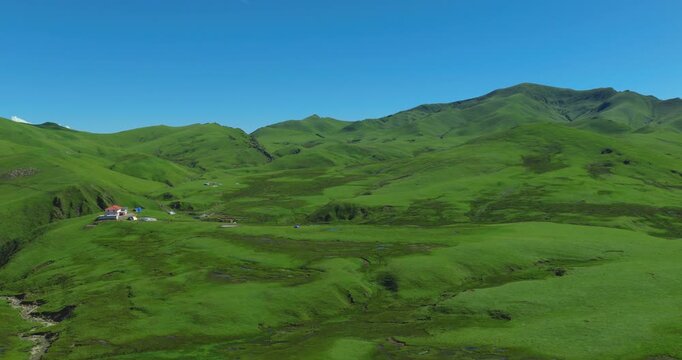 Badimalika region, Bajura, Nepal, drone shot of a small shelter building and tents perched on a lush green mountain ridge in Triveni Patan, surrounded by rolling alpine hills under a blue sky
