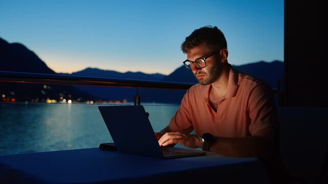 Focused freelance professional working on laptop at twilight balcony overlooking tranquil sea and mountains, blending remote work productivity with peaceful evening ambiance