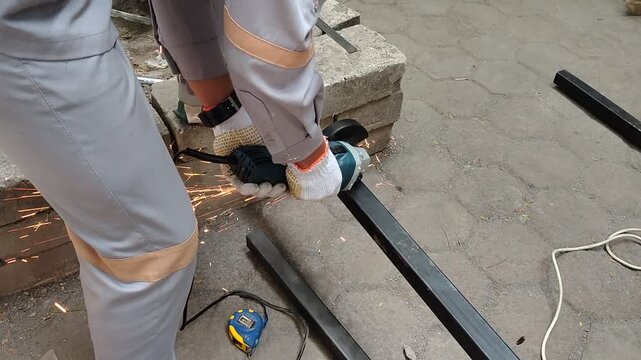 A worker in a grey uniform and safety gloves uses an angle grinder to cut a black square metal tube outdoors, creating bright flying sparks.