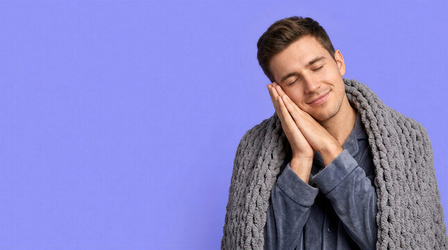 Relaxed young man miming sleep under a chunky gray blanket against a purple backdrop