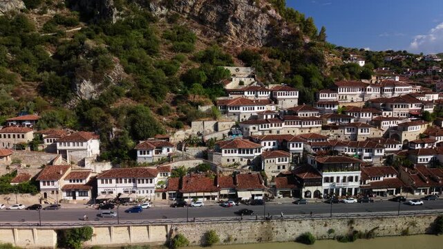 Berat Albania displays rows of traditional white houses across steep hillside above stone bridge and river. Historic town architecture and mountain greenery shape this bright urban panorama.