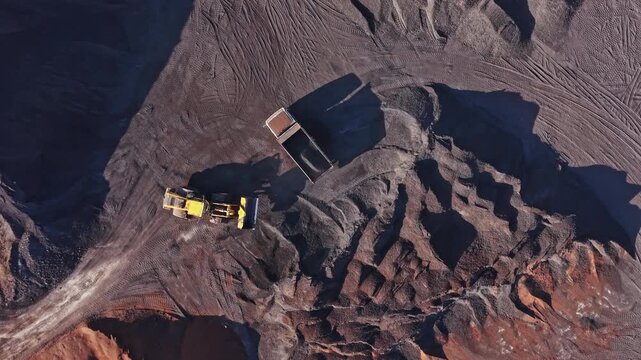An excavator loads a haul truck on a terraced mining site, with exposed rock and deep shadows across the pit during daylight.