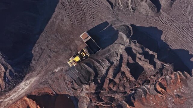An excavator and haul truck operate on a road cut into an open pit mine, with exposed rock and deep shadows across the site.