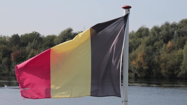 Belgium flag flying on a sunny day with Meuse river and wooded hill background at Dutch Belgium border town of Eijsden
