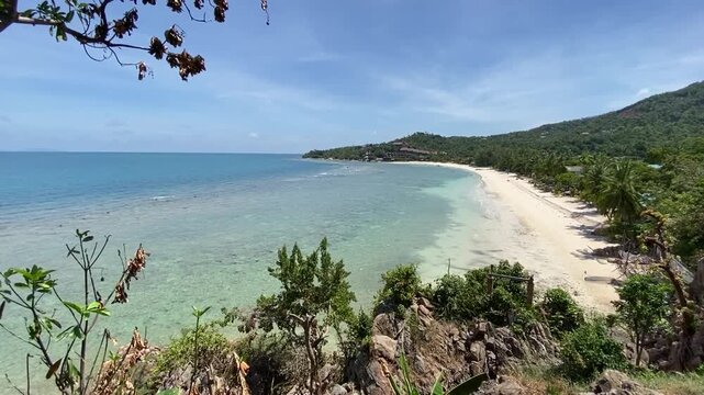 Aerial drone view of a curved white sandy beach bordered by turquoise coastal waters and rocky shores with lush green hills and scattered trees in Tri Trang Beach Phuket Thailand
