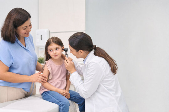Female Pediatrician Examining Little Girl Ear with Otoscope with Mother Supporting in Clinic with Copy Space
