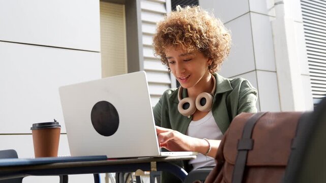 Young curly haired student typing on a laptop and studying online while sitting at an outdoor table of a university campus cafe, showing the modern concept of remote work and e learning