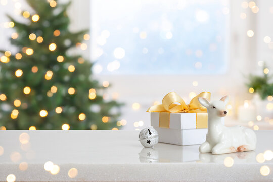 Festive Christmas background with white gift box, silver jingle bell and ceramic deer against blurred holiday tree interior