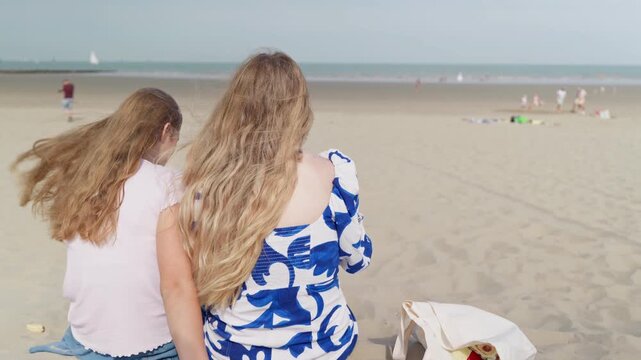 Unrecognizable mother and daughter are sitting on the beach slow motion view from the back the blurred sea in the background