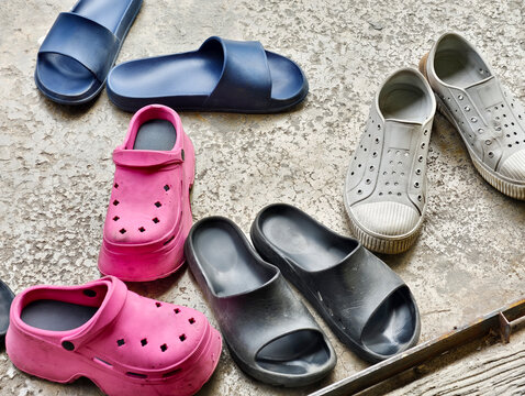 A top-down view of different pairs of casual rubber footwear, including pink clogs, slides, and slip-on shoes on a concrete floor. Ideal for daily life, family, and home entry concepts.