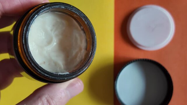 Hand holding an open amber jar to display a smooth cosmetic cream formulation.