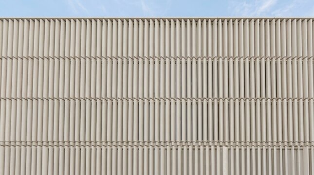 Contemporary building facade clad with vertical glazed ceramic louvers in light beige, creating a rhythmic texture under blue sky - Ventilated facade cladding and sun shading in modern architecture