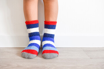 Child legs wearing colorful striped plush cozy warm socks standing on light wooden floor against white wall background indoor at home © Gulmira Uatkhanova