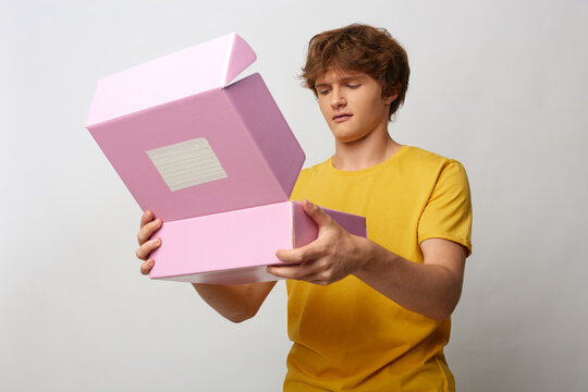 Young man in yellow tshirt looks with doubt into an open pink parcel box. His hesitant brow shows uncertainty. Concept of mail order and online shopping.