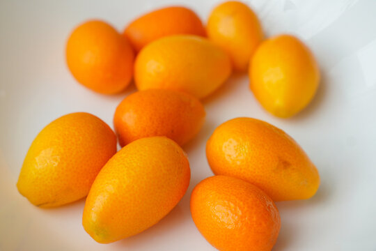 Several fresh kumquat fruits with bright orange skin arranged against a clean white background. These small citrus fruits feature an oval shape, vibrant color, and smooth, glossy surface. 