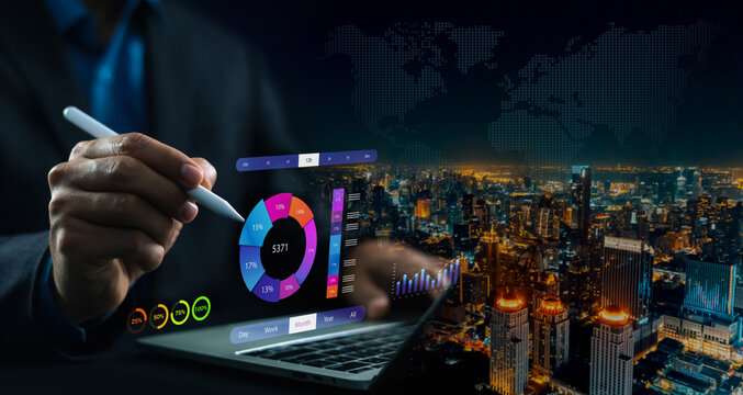 Businessman using a stylus to analyze financial data charts and statistics on a digital screen with a laptop and night city background. Concept of corporate business growth, analytics, and investment.