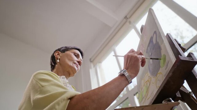 Senior Caucasian woman painting canvas on easel from low angle in art class. Footage suitable for promoting retirement leisure, creative education, and art therapy
