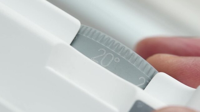 A hand rotates the gray thermostat wheel of a white electric heater from 14 to 26 degrees Celsius.